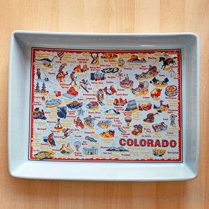 May include: A rectangular, light blue ceramic serving tray with a colorful map of Colorado. The map features illustrations of landmarks and towns, with the word "COLORADO" prominently displayed.