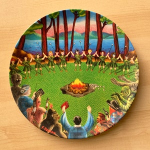 May include: A round ceramic plate with a colorful illustration of a group of people gathered around a campfire. The people are wearing colorful clothing and are holding sticks with marshmallows on them. The plate has a green background with trees and a lake in the background. The plate is signed "F. Pagow" in the bottom right corner.