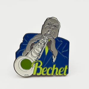 Pode incluir: Um broche de esmalte metálico com uma ilustração estilizada de um músico tocando saxofone. A figura tem pele cinza e veste um terno azul escuro. A palavra Bechet está escrita em letra cursiva verde-limão na base do saxofone.