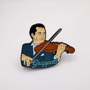 May include: A metal enamel pin featuring a stylized illustration of a musician playing a brown violin. The figure wears a dark blue suit jacket and white shirt. The name Grappelli is written in light blue script at the bottom of the pin.