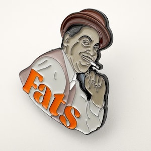 Peut inclure: Une broche en émail métallique représentant la caricature d'un homme portant un chapeau fedora marron et une chemise blanche. L'homme a une cigarette à la bouche et la main près du menton. Le mot Fats est écrit en grandes lettres orange.