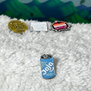 May include: A blue and white enamel pin of a soda can with the word "Joja" and "Cola" printed on it.