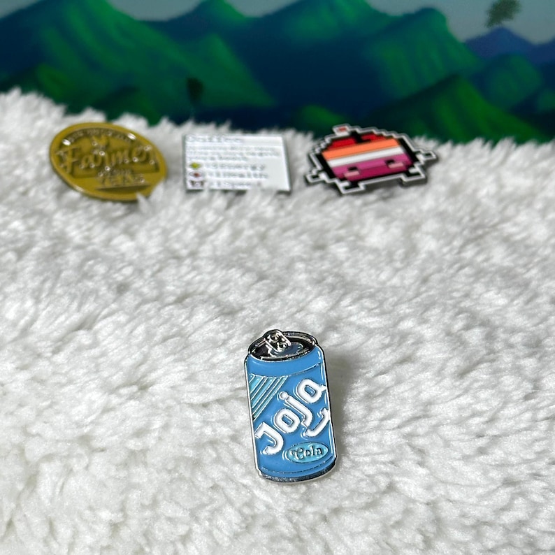 May include: A blue and white enamel pin of a soda can with the word "Joja" and "Cola" printed on it.