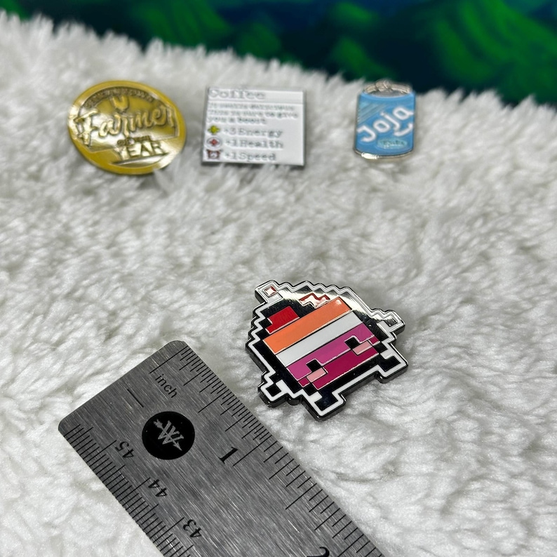 May include: A pixelated heart-shaped pin with a red, orange, white, and pink color scheme. The pin is on a white fuzzy surface with a ruler in the foreground.