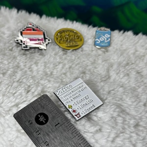 May include: Three enamel pins featuring a coffee cup, a can of Joja Cola, and a farmer's market logo. The coffee pin has text that reads "Coffee It smells delicious This is sure to give you a boost ++3 Energy +1 Health +1 Speed".