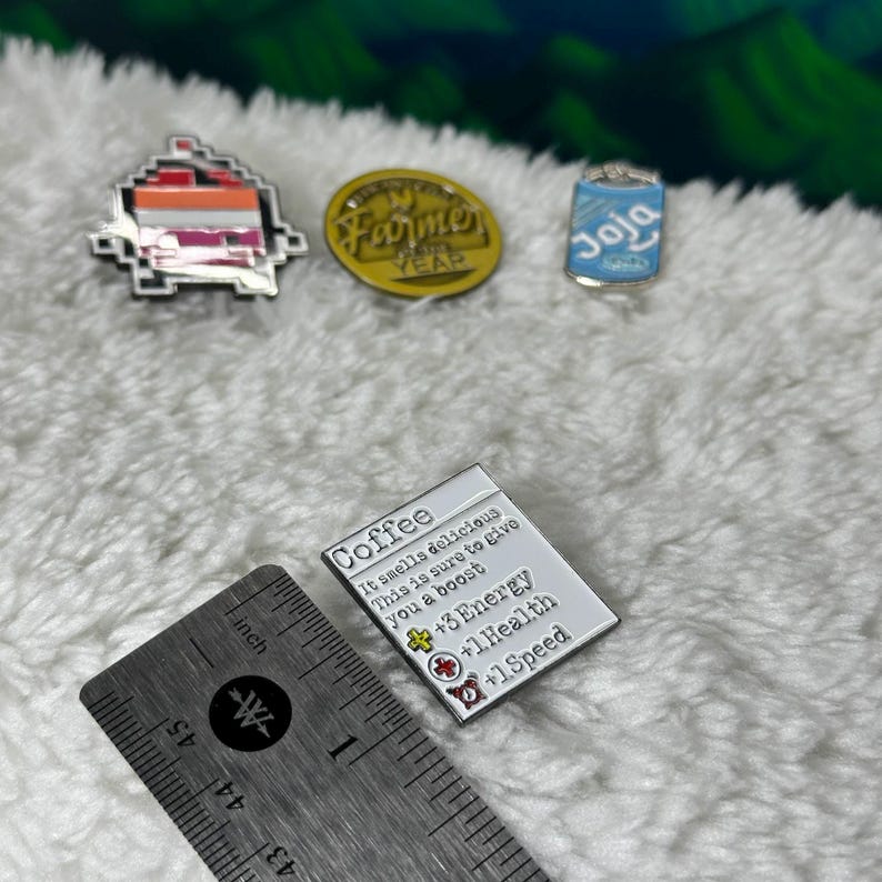 May include: Three enamel pins featuring a coffee cup, a can of Joja Cola, and a farmer's market logo. The coffee pin has text that reads "Coffee It smells delicious This is sure to give you a boost ++3 Energy +1 Health +1 Speed".