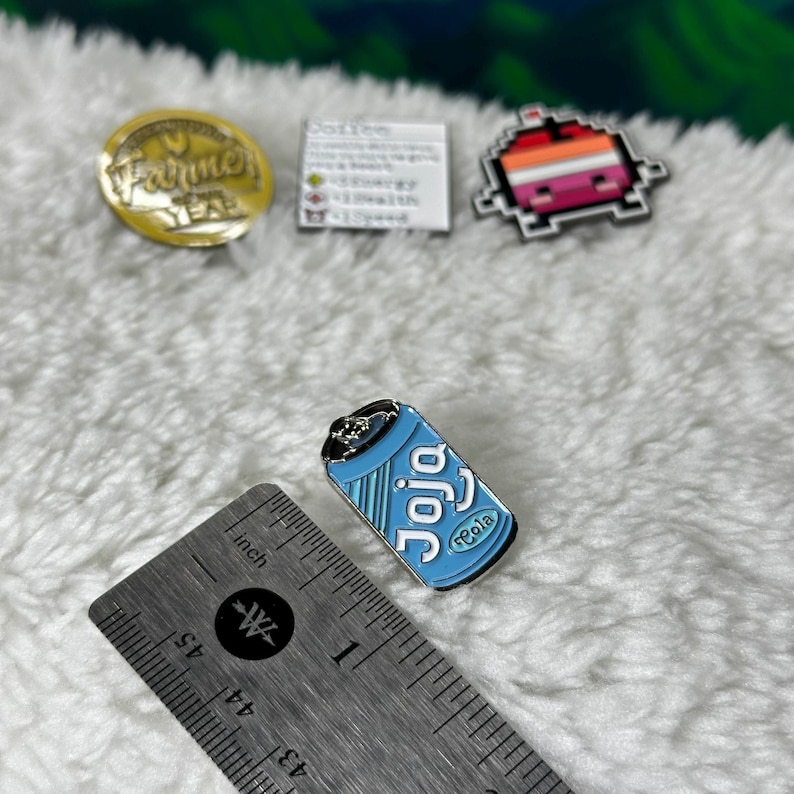 May include: A blue and white enamel pin of a can of soda with the word "JoJo" on it. The can is sitting on a ruler with inch markings.