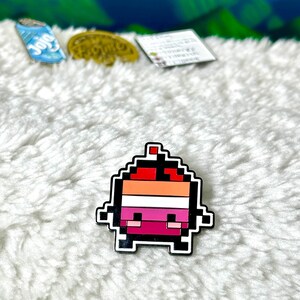 May include: A pixelated character pin with a red, orange, white, and pink color scheme. The character has a small red hat and a smiling face.