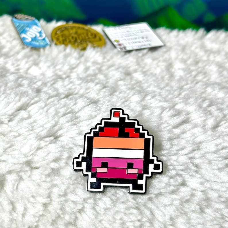 May include: A pixelated character pin with a red, orange, white, and pink color scheme. The character has a small red hat and a smiling face.