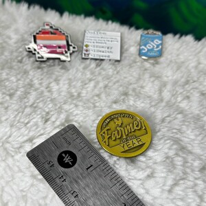 May include: Four enamel pins: a pixelated pink, orange, and white character, a blue can with the word "Joja" on it, a white card with the text "Dot.exe" and a gold pin with a chicken and the text "Pelican Town Farmer of the Year".