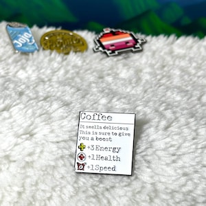 May include: White enamel pin with a coffee cup graphic and text that reads "Coffee" and "It smells delicious. This is sure to give you a boost. +3 Energy +1 Health +1 Speed".
