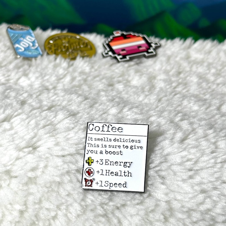 May include: White enamel pin with a coffee cup graphic and text that reads "Coffee" and "It smells delicious. This is sure to give you a boost. +3 Energy +1 Health +1 Speed".