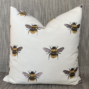 May include: White throw pillow with an embroidered pattern of bumblebees. The bees are yellow and black with brown wings.