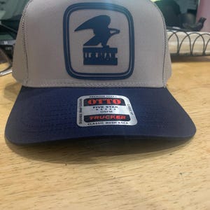 Op de afbeelding: Een grijze en marineblauwe truckerpet met een mesh achterkant. De voorkant heeft een marineblauwe vierkant met het logo van de Amerikaanse postdienst. De pet heeft een druksluiting en een klassieke pasvorm.