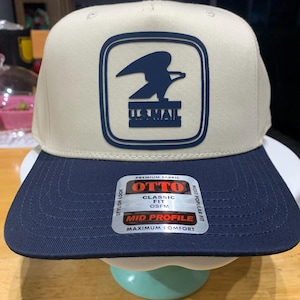 Könnte beinhalten: Beige und marineblaue Baseballkappe mit flachem Schirm. Die Kappe hat einen marineblauen quadratischen Aufnäher mit dem US Mail-Logo. Auf dem Schirm befindet sich ein Aufkleber mit der Aufschrift "OTTO" und "MID PROFILE".