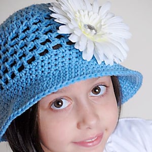 May include: A young girl wearing a light blue crocheted sun hat with a white flower on top. The hat has a wide brim and a crocheted design.