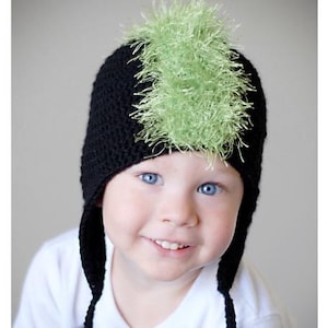 May include: A child wearing a black knit hat with a green fuzzy mohawk. The hat has ear flaps and is crocheted.