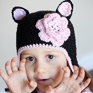 Puede incluir: Un gorro de crochet negro con orejas rosas y una flor rosa en la parte superior. El gorro tiene un borde rosa alrededor del borde y una solapa rosa que cubre las orejas. Un niño pequeño lleva el gorro.
