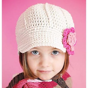 Peut inclure: Un chapeau blanc en crochet avec une fleur rose sur le côté. Le chapeau a une visière et est porté par une jeune fille aux cheveux bruns.