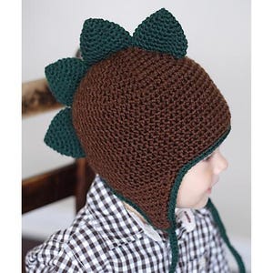 May include: A brown crocheted hat with green dinosaur spikes and ear flaps. The hat is being worn by a child.