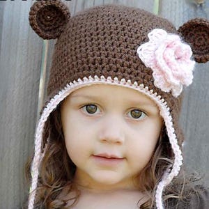 Może przedstawiać: Brązowy, dziergany na drutach kapelusz z uszami niedźwiedzia i różową, dzierganą na szydełku kwiatkiem. Kapelusz ma różową, dzierganą na szydełku lamówkę i sznurki. Małe dziecko nosi kapelusz.