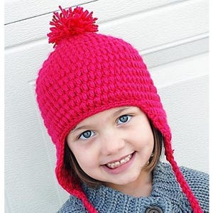 Puede incluir: Un gorro de punto de color rosa brillante con un pompón en la parte superior y lazos largos. El gorro lo lleva puesto un niño con el pelo castaño y los ojos azules.