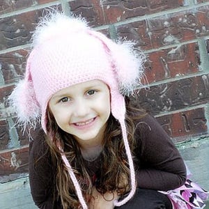 May include: A pink crocheted hat with white fluffy pom-poms and ear flaps. The hat has a long tie that hangs down the back of the neck.