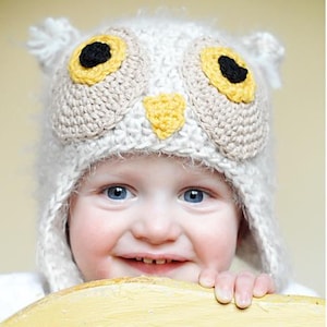 May include: A white crocheted owl hat with yellow and black felt accents. The hat has a fluffy texture and is worn by a child.