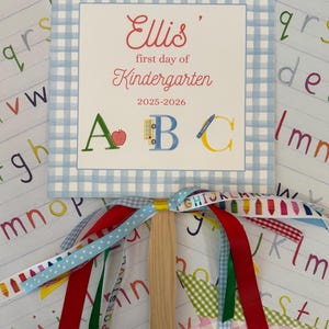 May include: A light blue and white gingham-patterned sign with the text "Ellis' first day of Kindergarten 2025-2026." The sign has a wooden handle and colorful ribbons attached. The letters A, B, and C are displayed with illustrations.