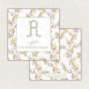 Op de afbeelding: Twee vierkante cadeaulabels met een bloemenontwerp. De labels hebben een lichtroze achtergrond met een patroon van bloeiende takken in roze, geel en groen. Eén label heeft de groene initiaal "R" en de tekst "a gift from THE ROBERTS FAMILY."
