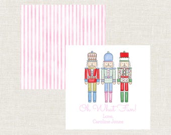 Può includere: Un biglietto quadrato con tre soldati schiaccianoci illustrati in uniformi rosse, blu e verdi. Il biglietto presenta il testo "Oh What Fun!" e "Love, Caroline Jones" in rosa. Dietro c'è un biglietto a righe rosa e bianche.