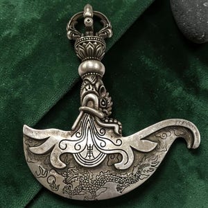 May include: A silver-colored ritual knife with intricate carvings. The handle features a floral design and a detailed face. The curved blade is decorated with a dragon and other ornate patterns. The knife is displayed on a green surface.