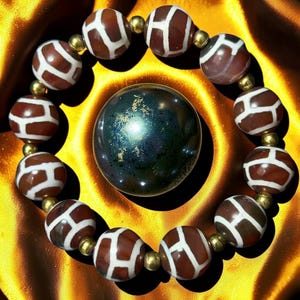 Op de afbeelding: Een kralenarmband met bruine en witte kralen met patronen en goudkleurige afstandhouders omsluit een donkere, iriserende bol. De kralen van de armband hebben een uniek ontwerp en de bol reflecteert licht.