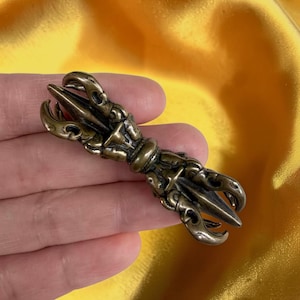 Puede incluir: Un vajra de color bronce, un objeto ritual con un punto central y extremos en forma de garra. El vajra se sostiene en una mano sobre un fondo amarillo. El objeto tiene detalles intrincados y un acabado pulido.