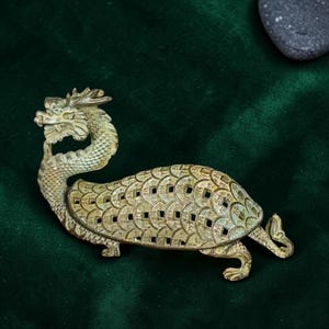 Peut inclure: Figurine dragon-tortue de couleur bronze. La tête de dragon est ornée d'une moustache bouclée et d'une petite corne. La carapace de la tortue est faite de formes de pièces de monnaie reliées. Sur fond vert foncé.