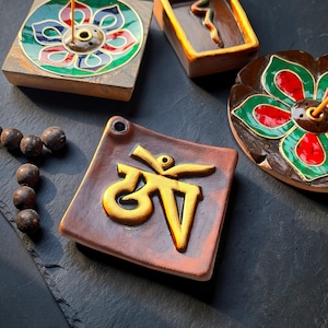 May include: A collection of incense holders in various shapes and designs. One is square with a gold Om symbol, another is a flower shape with red and green accents. Other holders are rectangular with brown and gold colors.