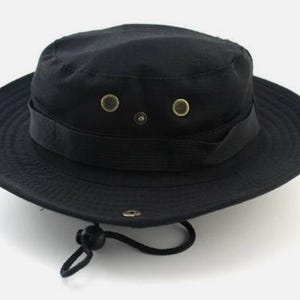 May include: A solid black bucket hat featuring a wide brim, a flat crown, and a fabric band around the base. The hat includes two metallic ventilation eyelets on each side, a small metal snap button on the brim, and an adjustable black chin cord.