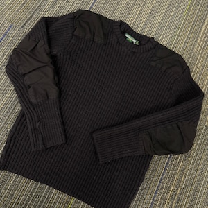 May include: A black long-sleeve sweater featuring a ribbed knit texture, a crew neckline, and solid black fabric patches reinforced on the shoulders and elbows. The garment has a straight hemline and a label visible inside the collar area.