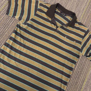 May include: A short-sleeved polo shirt featuring horizontal stripes in olive green, dark charcoal, and thin bright turquoise lines. The shirt includes a solid dark charcoal collar, a two-button placket, and a single chest pocket with metal rivets.