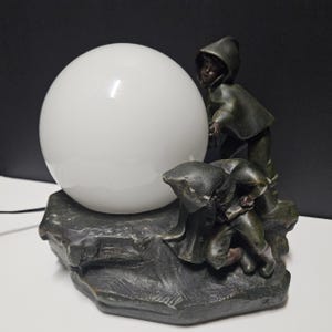 May include: A decorative lamp featuring a large white globe and two figures in hooded cloaks. The figures appear to be interacting with the globe, which sits atop a dark, textured base. The lamp has a vintage aesthetic.