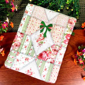 May include: A patchwork fabric case with a floral pattern featuring pink roses and green accents. The case has a green bow applique and a small red bow. The case is rectangular and has a zipper closure.