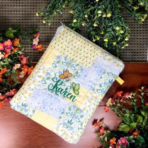 May include: A quilted, zippered case with a patchwork design in yellow, white, and blue floral patterns. The case features an embroidered butterfly and the name "Karen" in green thread. A yellow zipper pull is visible.