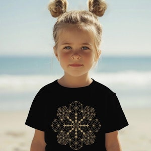 May include: A young person wearing a black t-shirt with a gold geometric design. The design is a mandala with a flower of life pattern. The person has their hair pulled back in two buns.