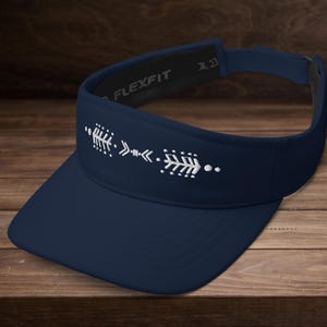 Puede incluir: Visera azul marino con un diseño de flecha blanca. La visera tiene una visera curva y una correa ajustable. El texto "FLEXFIT" es visible en el interior de la visera.