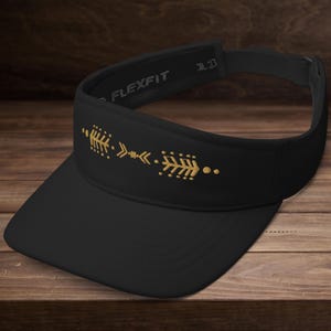 Puede incluir: Visera Flexfit negra con un diseño de flecha dorada. La visera tiene una visera curva y una correa ajustable. El diseño de la flecha está formado por pequeñas formas geométricas y puntos. La visera está sobre una superficie de madera.