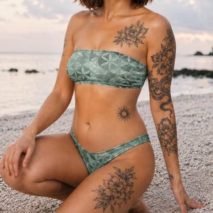 Könnte beinhalten: Ein salbeigrüner trägerloser Bikini mit geometrischem Blumenmuster. Das Oberteil ist im Bandeau-Stil gehalten und das Unterteil ist ein hoch geschnittener Bikini-Slip. Der Stoff hat ein dezentes, sich wiederholendes Muster aus Linien.