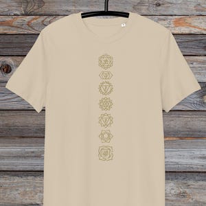 May include: A beige t-shirt with a vertical line of seven gold chakra symbols printed on the front. The symbols are in a line down the centre of the shirt.
