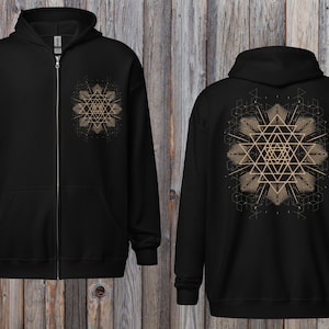 May include: Black zip-up hoodie with a geometric design on the front and back. The design is a gold-colored, symmetrical pattern. The hoodie has a hood and a front pocket. The background is a wooden surface.