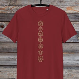 May include: A maroon t-shirt with a vertical row of seven gold chakra symbols printed on the front. The symbols are in a line down the centre of the shirt.