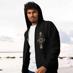 May include: Black zip-up hoodie with a gold geometric design on the front. The hoodie has a hood and a white t-shirt underneath. The person is standing in front of a beach.